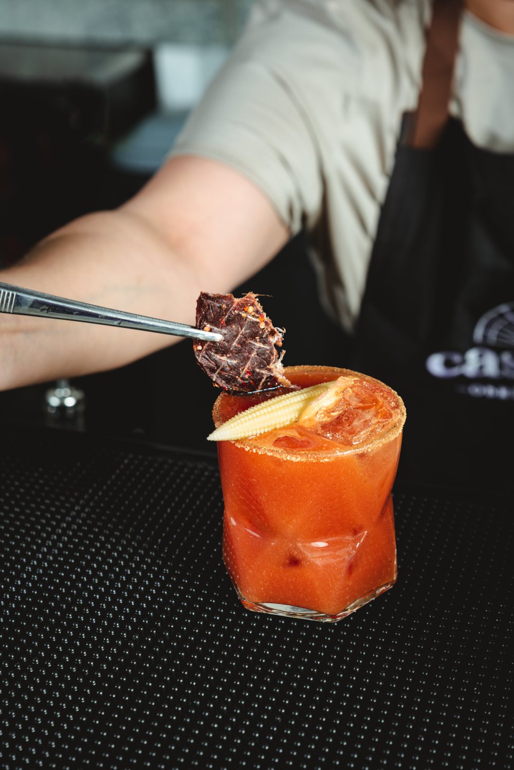 Bebida preparada de la barra con escarcha, tomate y elote baby, terminada con una lámina de carne seca.
