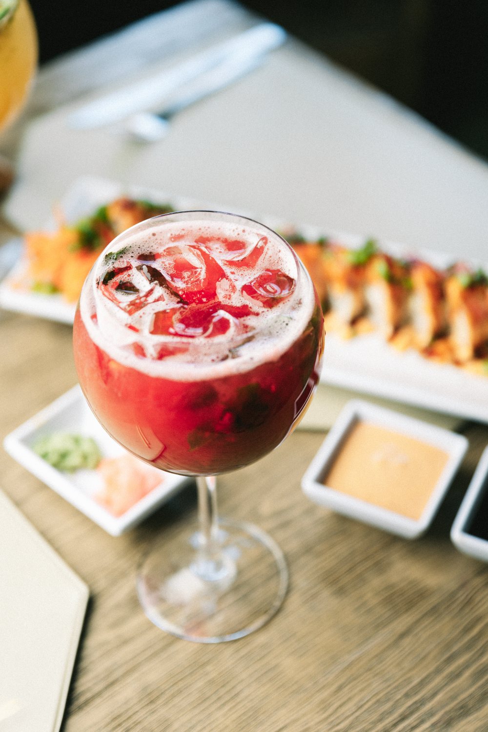 Trago de frutos rojos en copa balón, con hielo y hierbabuena, servido junto a un roll de sushi.