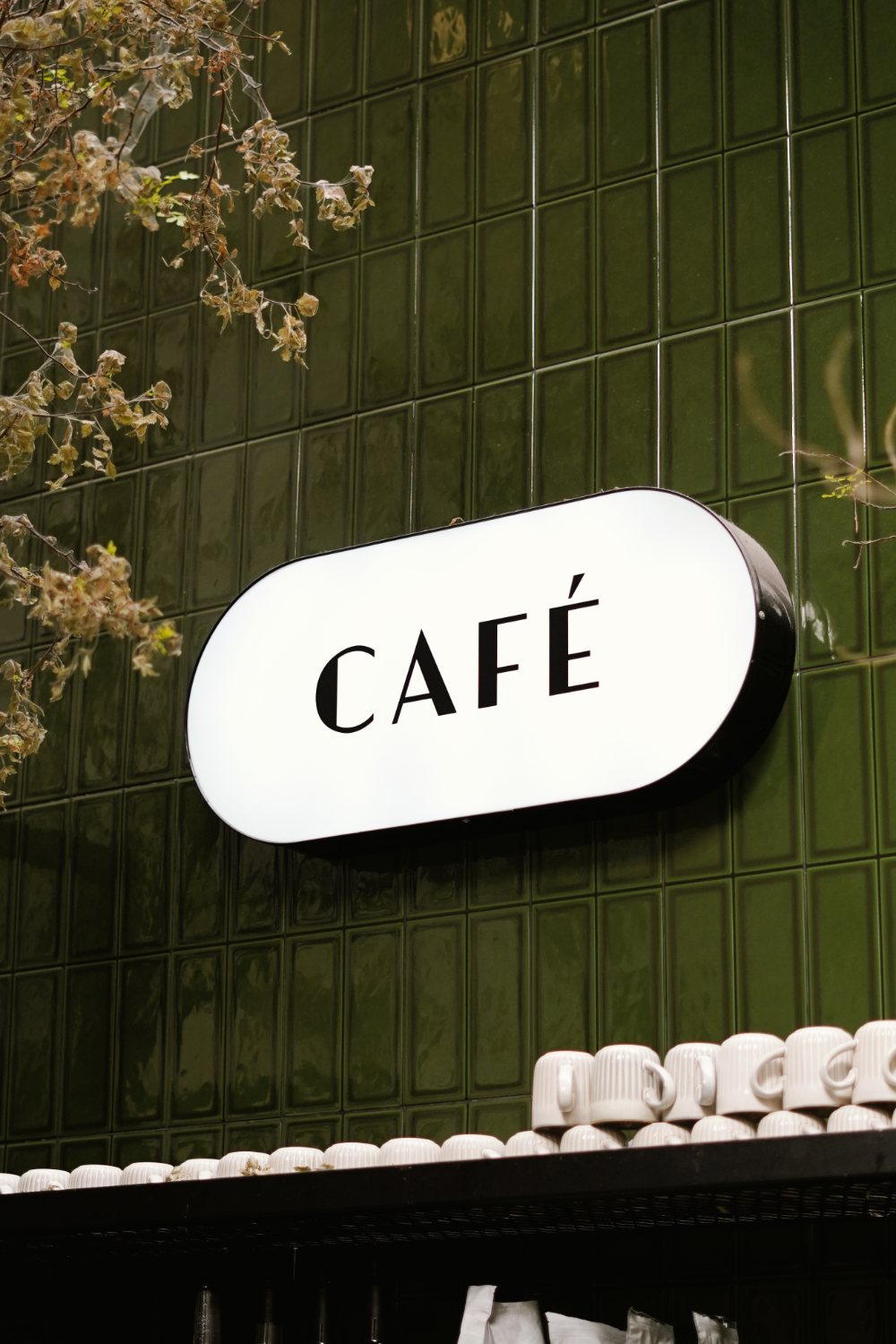 Letrero luminoso con la palabra Café sobre muro de azulejo verde, con las tazas apiladas de la barra.