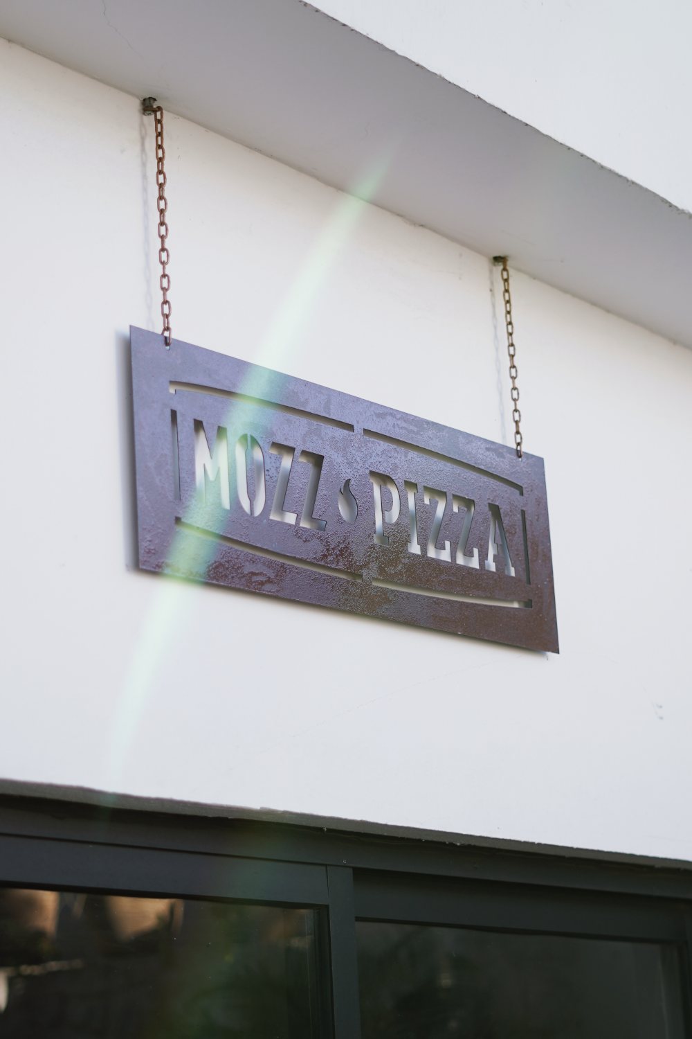 Letrero de acero de Mozz Pizza colgado de cadenas sobre la ventana.