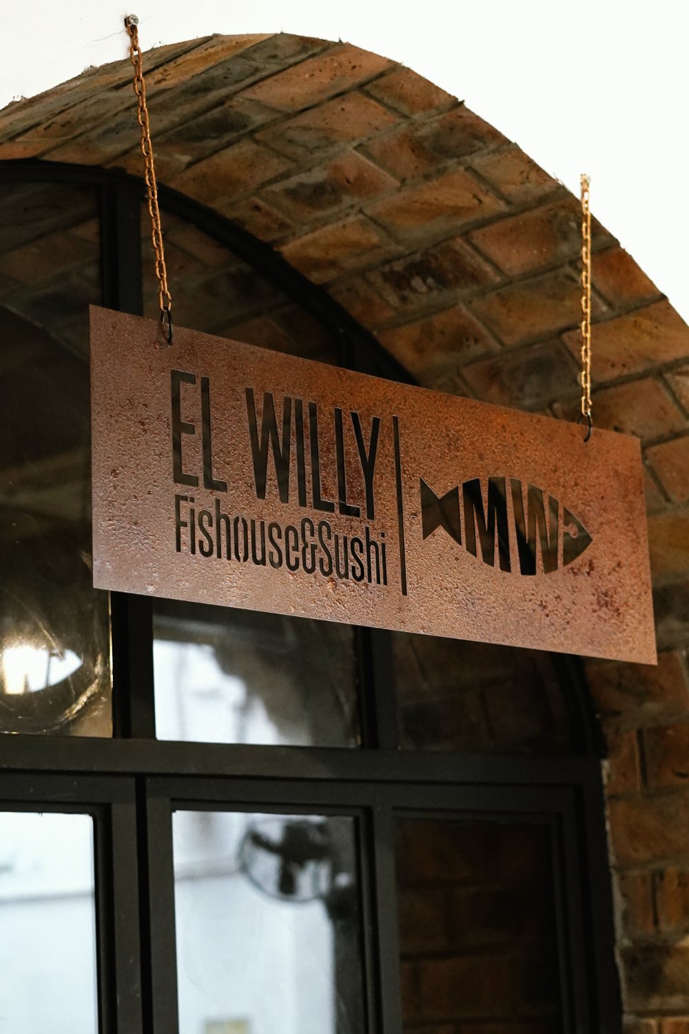 Letrero de acero cortado de El Willy Fishouse & Sushi, colgado de cadenas bajo un arco de ladrillo.