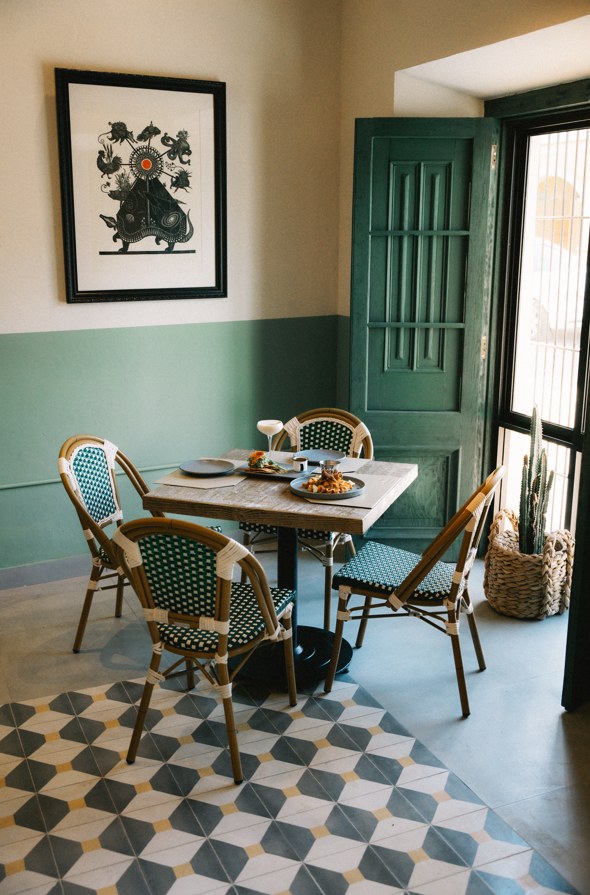 Mesa de bistró servida junto a la puerta verde, con zoclo salvia, piso de pasta y un grabado enmarcado en el muro.