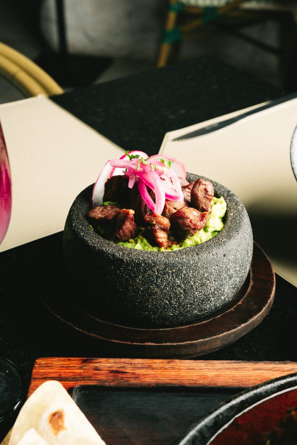 Molcajete de piedra con guacamole, carne asada y cebolla morada encurtida, sobre base de madera.